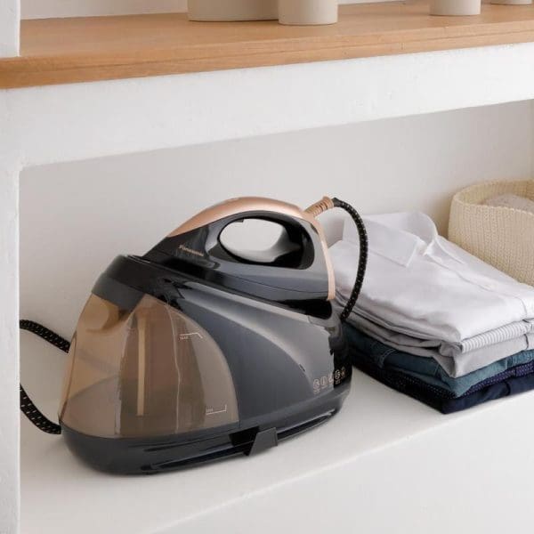 Panasonic NI-GT500 anti-calc steam generator iron in grey and rose gold on white surface with folded clothes and laundry basket nearby.