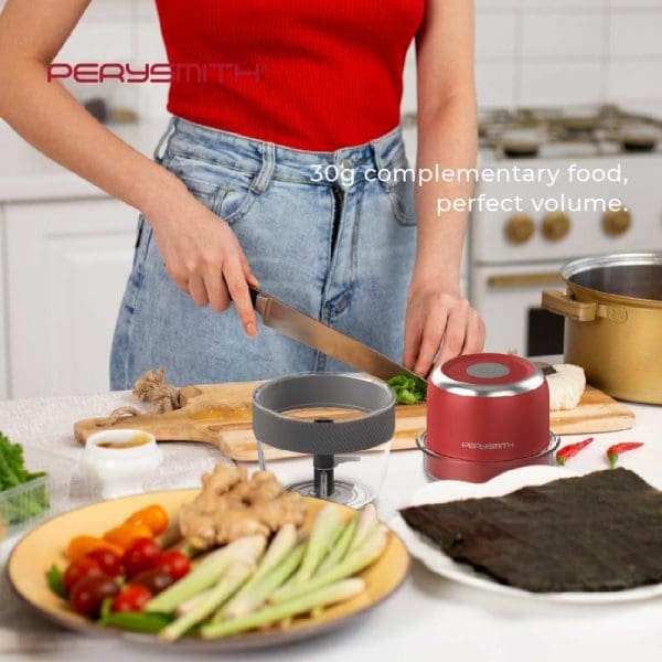 Woman in red top and jeans using PerySmith Mini Food Chopper PS250 in modern kitchen with fresh vegetables, cutting board, and cooking ingredients on counter.