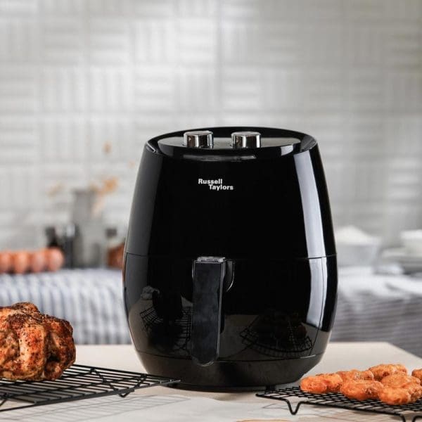 Russell Taylors AF-34 XL 4.8L black air fryer on kitchen counter with cooked chicken and fried food on cooling racks nearby.