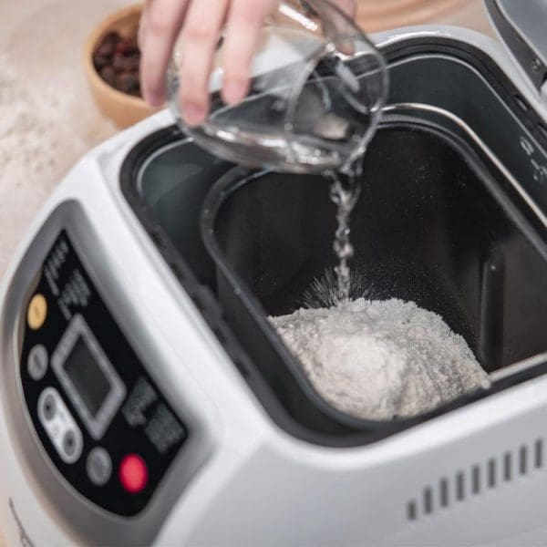 Russell Taylors Bread Maker BM-10 with hand pouring water into flour in black non-stick pan, digital control panel visible, kitchen countertop setting.
