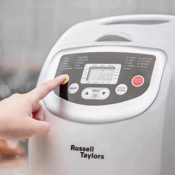 Russell Taylors BM-10 bread maker with digital display showing 13:00 timer, control panel with various bread settings, and hand pressing button on white appliance.