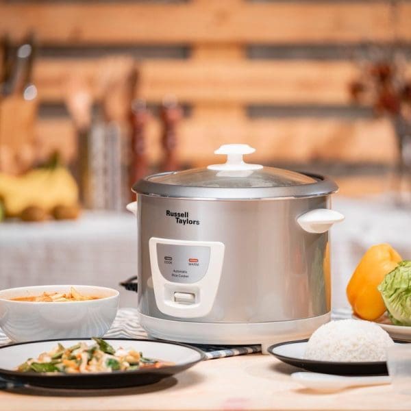 Russell Taylors 1.8L stainless steel rice cooker with glass lid on kitchen counter, surrounded by cooked rice, vegetables, and fresh produce.