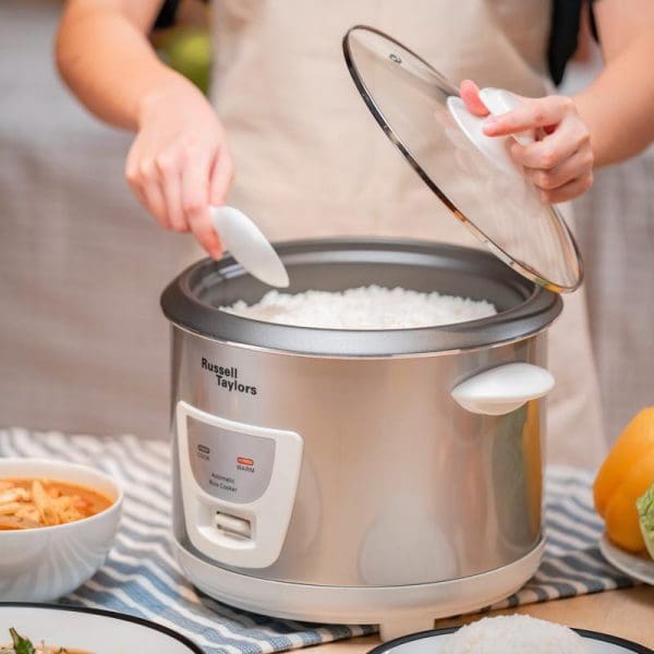 Russell Taylors ERC-18 1.8L rice cooker with steam rack, person lifting glass lid revealing cooked white rice inside stainless steel pot.