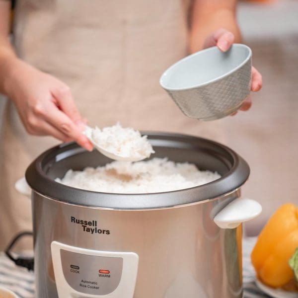 Person using Russell Taylors ERC-18 1.8L rice cooker, serving cooked white rice with wooden spoon while holding ceramic bowl in kitchen setting.