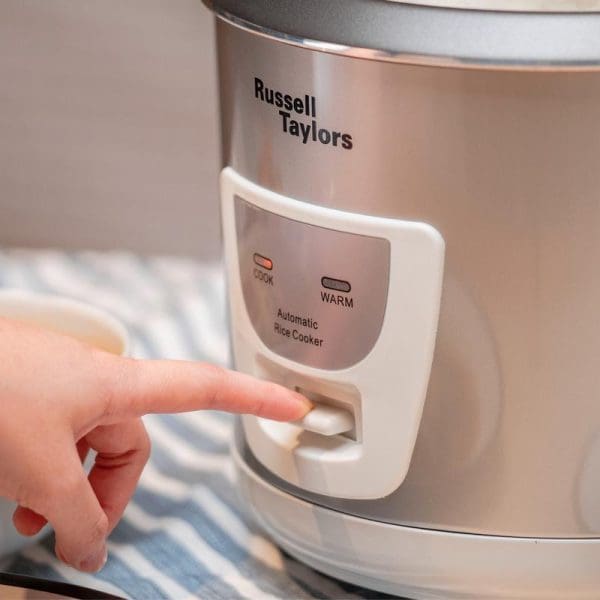 Russell Taylors automatic rice cooker with hand pressing white switch button, showing COOK and WARM settings on rose gold appliance.
