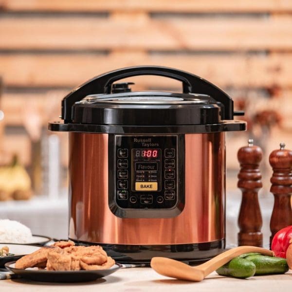 Russell Taylors PC-12 copper-colored electric pressure cooker with digital display on kitchen counter surrounded by vegetables, wooden utensils, and salt and pepper shakers.