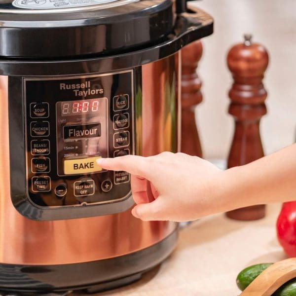 Russell Taylors electric pressure cooker with copper finish, digital display showing BAKE mode, hand pressing button, wooden pepper mills in background on kitchen counter.
