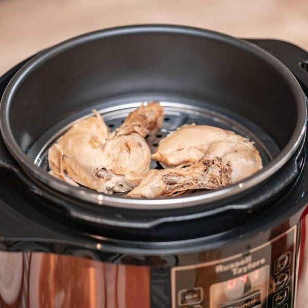 Russell Taylors PC-80 8L electric pressure cooker with stainless steel pot steaming chicken pieces on metal rack inside black cooking vessel.
