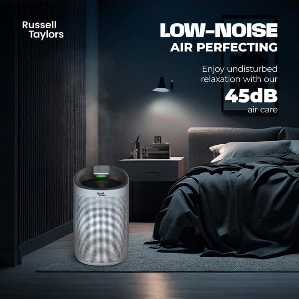 Russell Taylors white cylindrical air purifier in modern dark bedroom with unmade bed, highlighting 45dB low-noise operation for undisturbed relaxation and sleep.
