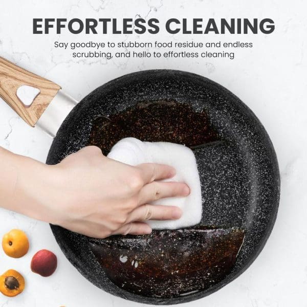Hand cleaning a black speckled non-stick frying pan with white cloth, demonstrating effortless cleaning with wooden handle and fresh fruits nearby on marble surface.