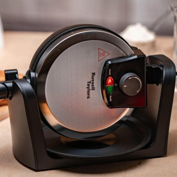 Russell Taylors WM-30 rotating waffle maker with black stand, stainless steel cooking plates, red and green indicator lights, and temperature control dial on kitchen counter.