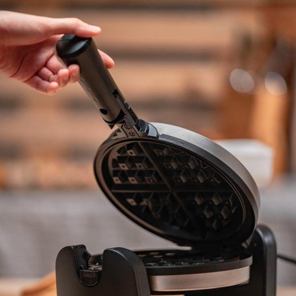 Russell Taylors Rotating Waffle Maker WM-30 with open lid showing waffle grid pattern, hand holding black handle, kitchen background