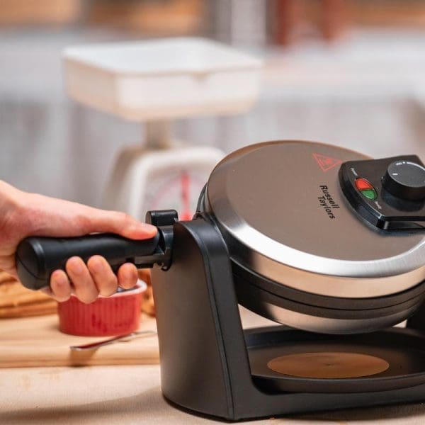 Russell Taylors WM-30 rotating waffle maker in black with control dial, showing hand holding handle and red ramekin on wooden kitchen counter.