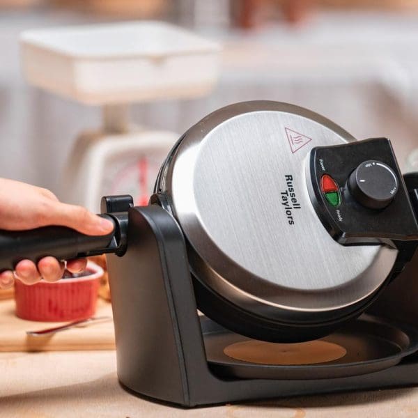 Russell Taylors WM-30 rotating waffle maker with stainless steel lid and black base, featuring temperature control dial and indicator lights, shown in kitchen setting.