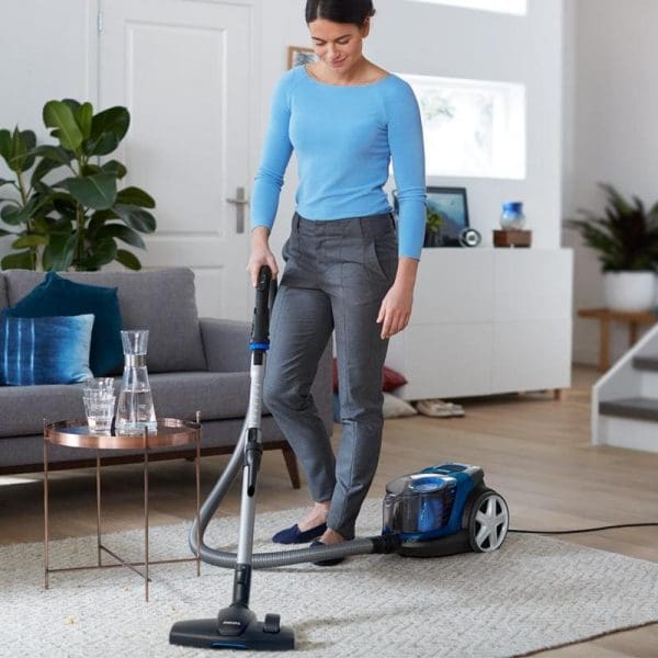 Wanita menggunakan Pembersih Vakum Tanpa Beg Philips FC9352 PowerPro Compact di ruang tamu moden dengan sofa kelabu dan tumbuhan hijau