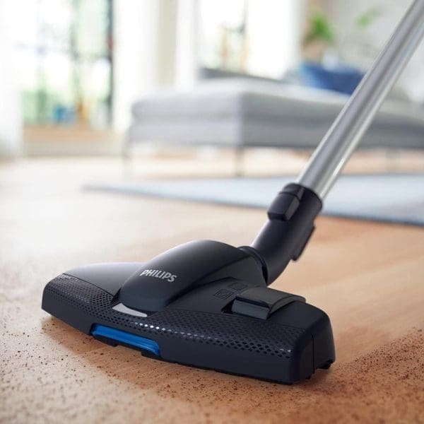 Philips FC9352 PowerPro Compact penyedut habuk tanpa beg sedang membersihkan permaidani coklat di ruang tamu dengan sofa kelabu di latar belakang