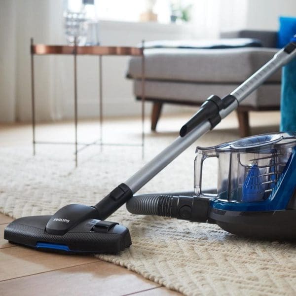 Philips FC9352 PowerPro Compact penyedut habuk tanpa beg sedang membersihkan permaidani berbulu di ruang tamu dengan sofa dan meja kaca di latar belakang.